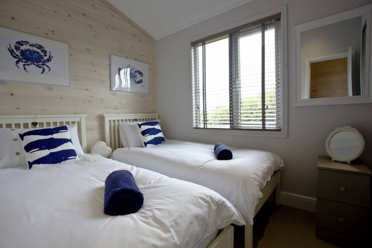 A bedroom with two beds, a window with blinds, a bedside table with a mirror, and artwork on the wall.