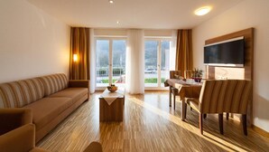 A modern living room with a sofa, a dining table, and chairs. There is a TV mounted on the wall and a window with curtains. - Hotel zum Anker (Neumagen-Dhron)
