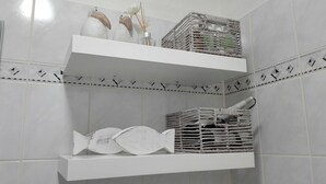 Une salle de bain avec des murs recouverts de carreaux blancs, deux étagères blanches et un poisson décoratif sur l’étagère du bas.