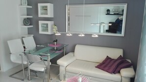 Un salon moderne avec une table à manger en verre, des chaises blanches, un canapé blanc agrémenté de coussins violets, un grand miroir et une petite plante en pot.