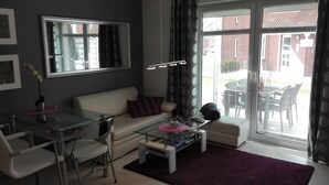 Un salon moderne avec une table à manger en verre, un canapé et une table basse. Une grande fenêtre, dotée de stores, offre une vue sur l’extérieur.