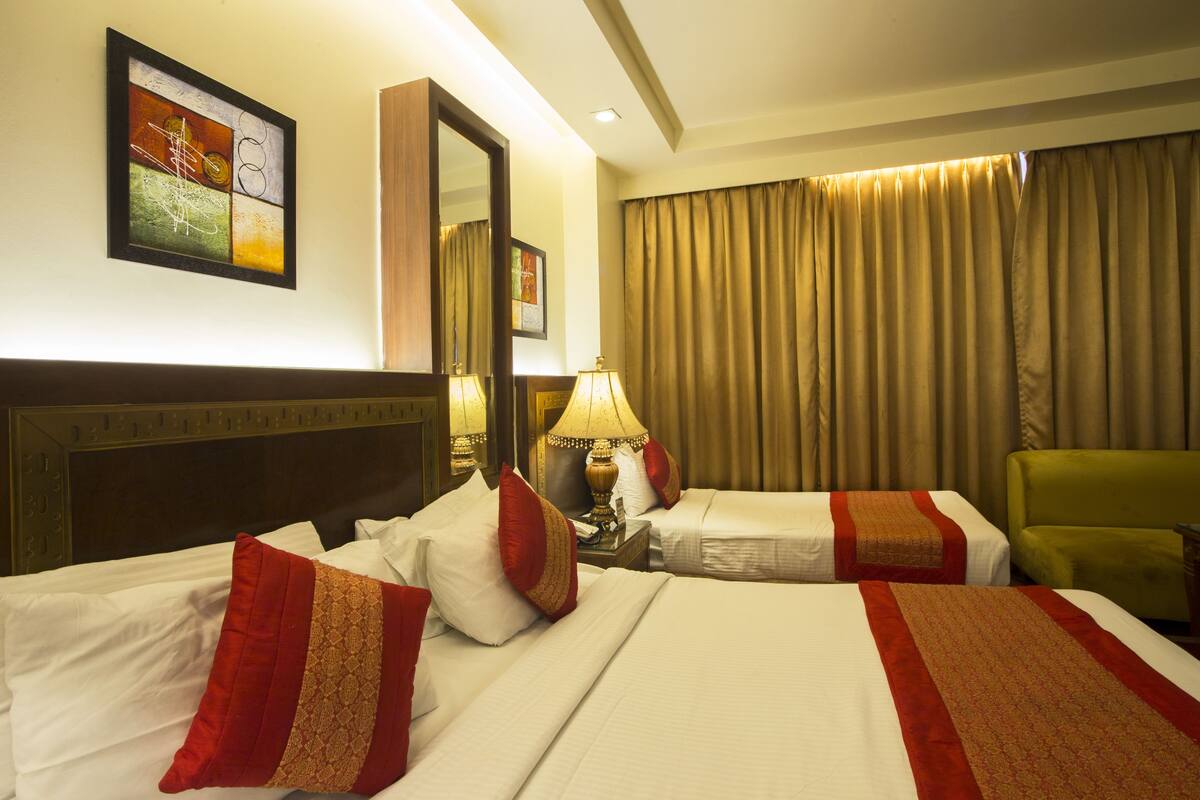 A hotel room with two beds, a green armchair, a mirror, and a painting on the wall.