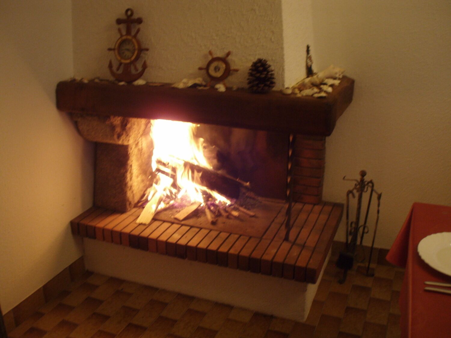 Une cheminée surmontée d’une étagère décorée d’objets marins, un sol à carreaux et une table avec une assiette et des baguettes.