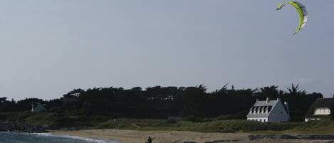Een strand met een persoon die kitesurft, huizen op de achtergrond en een heldere lucht.