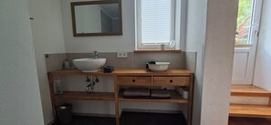 A bathroom with a white sink, a mirror, a window with blinds, a towel rack, and a wooden shelf.