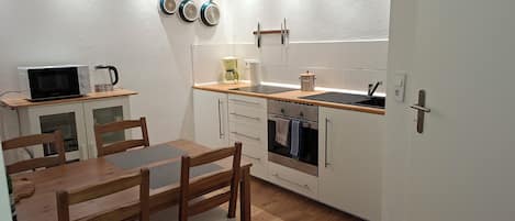 Una cocina compacta con microondas, hervidor y tetera sobre la encimera. Hay una mesa de comedor de madera con sillas.
