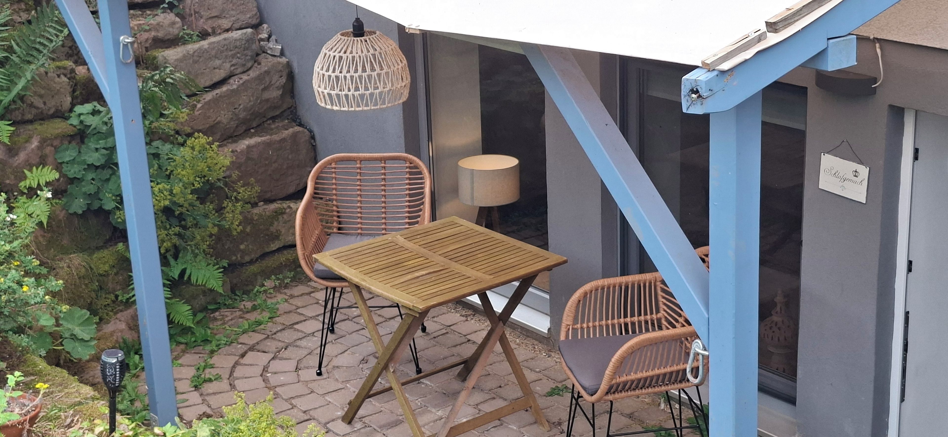 An outdoor seating area with wicker chairs and a wooden table, a hanging light fixture, and a stone wall.