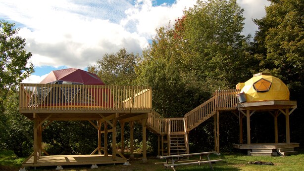 A wooden deck with a red and yellow tent structure, surrounded by trees and a clear sky. - Apple Dome at Cabot Shores Wilderness Resort (Englishtown)