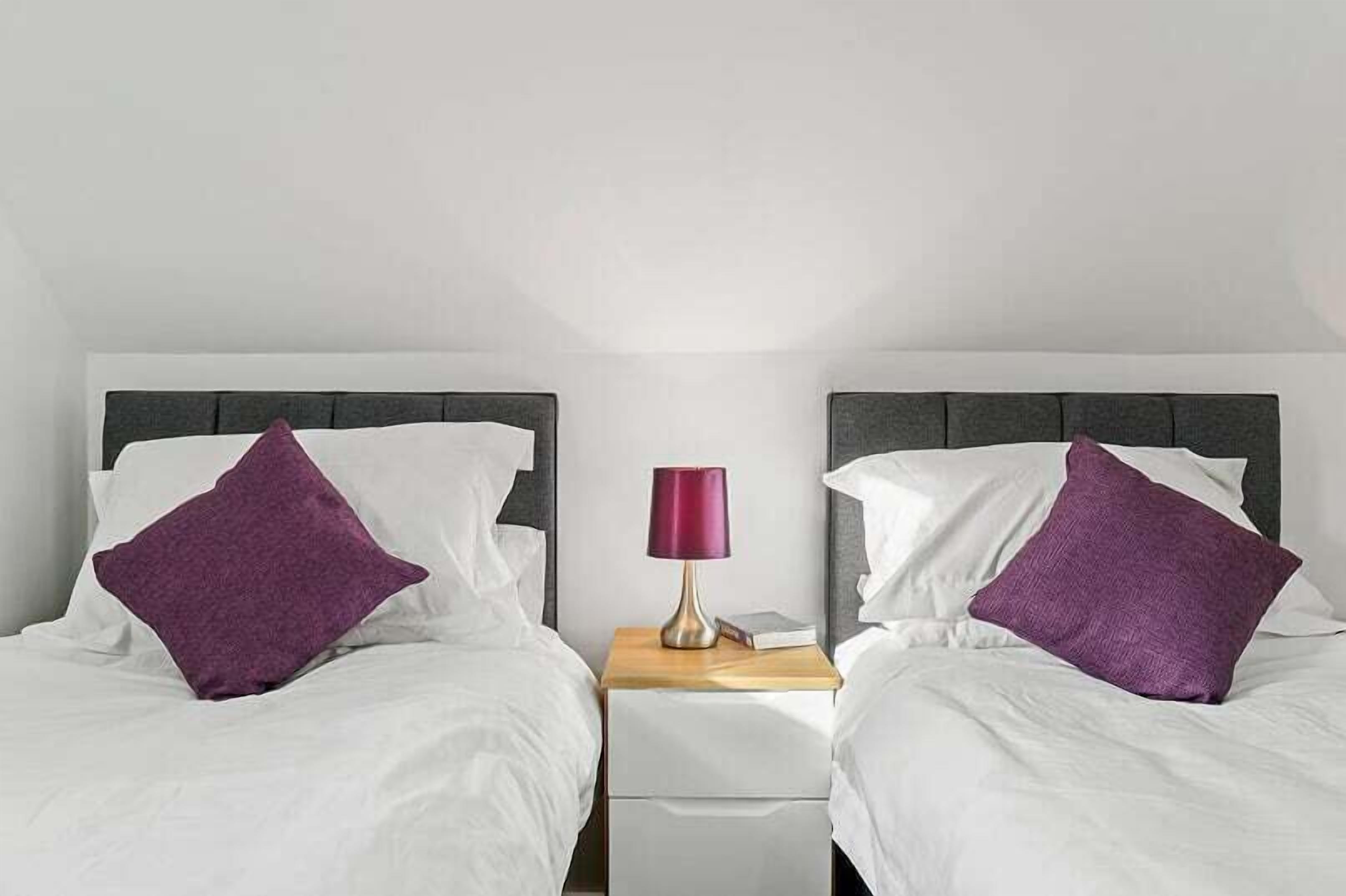 A twin room with two beds, each with white bedding and purple pillows, a nightstand with a lamp, and a small table between the beds.