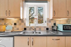 Una cocina con armarios de madera, un lavamanos doble, una tetera, un microondas y una ventana con vistas a árboles.