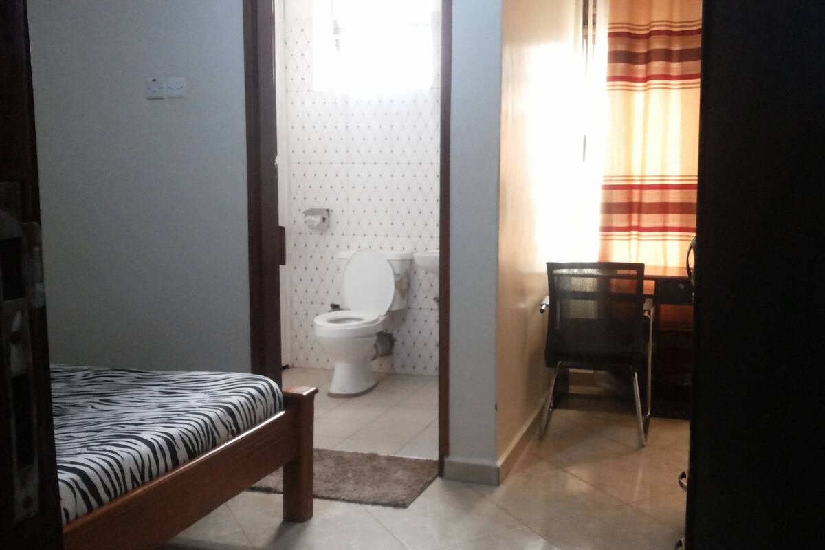 A bedroom with a bed, a chair, a desk, and a bathroom with a toilet and sink.