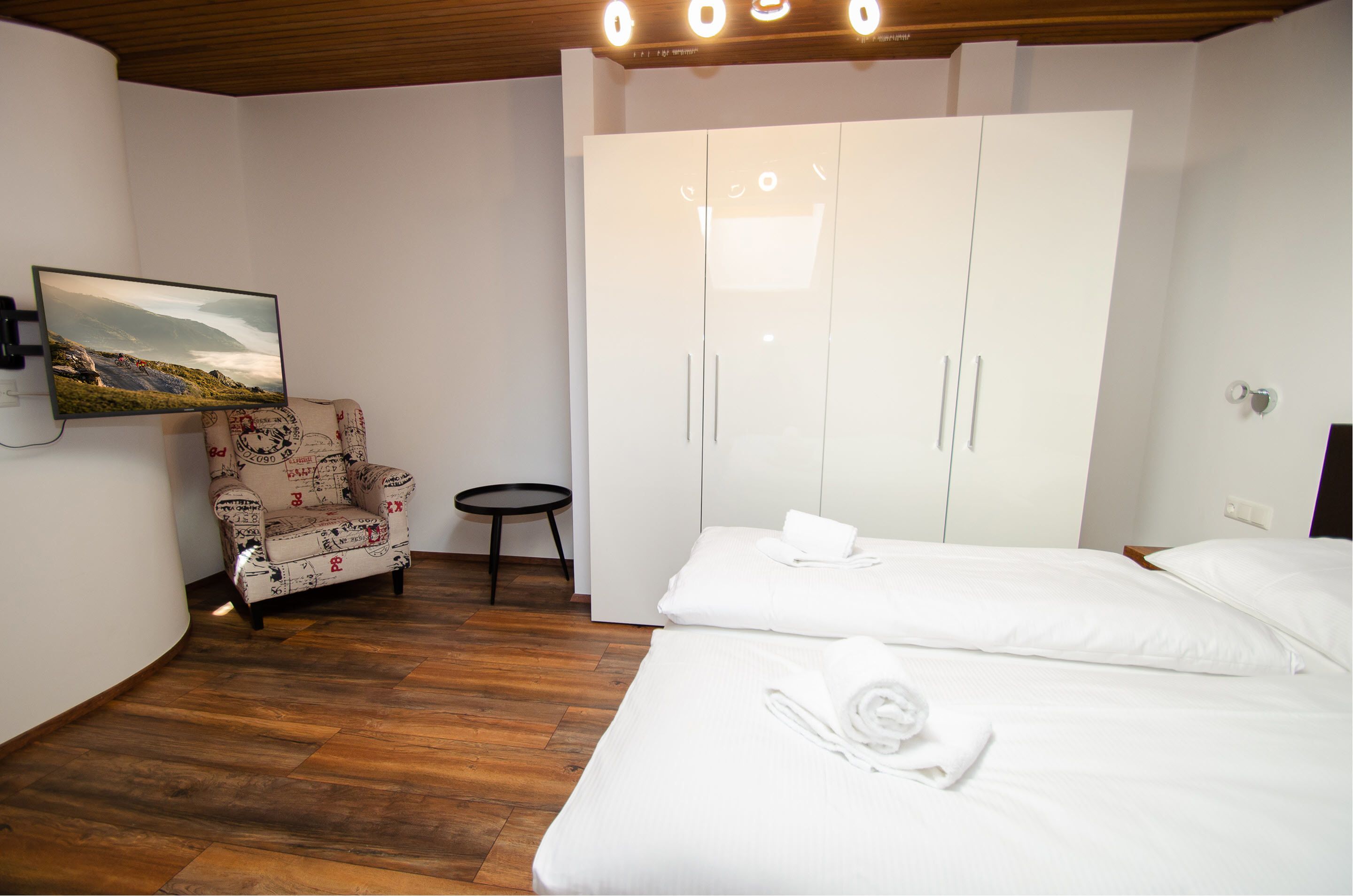A hotel room with a bed, a wardrobe, a chair, a small table, and a wall-mounted TV.