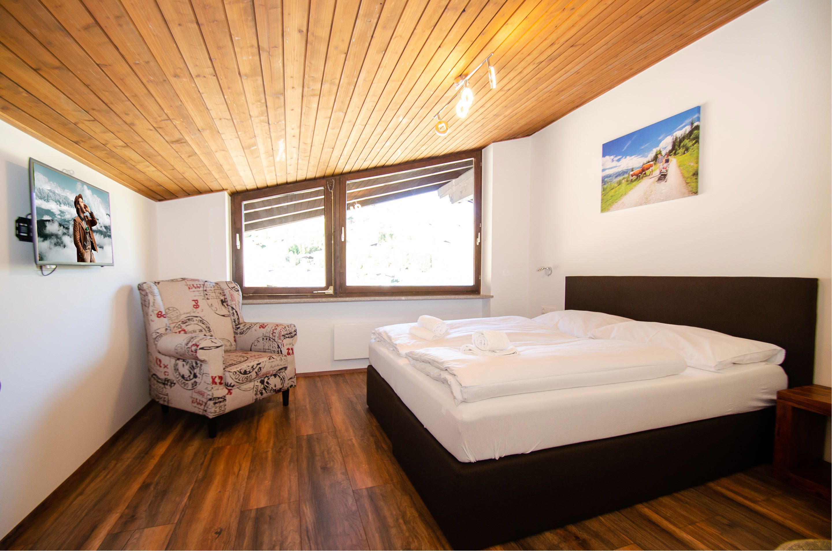 A bedroom with a wooden ceiling, a bed, a chair, and a window with a view of the countryside.