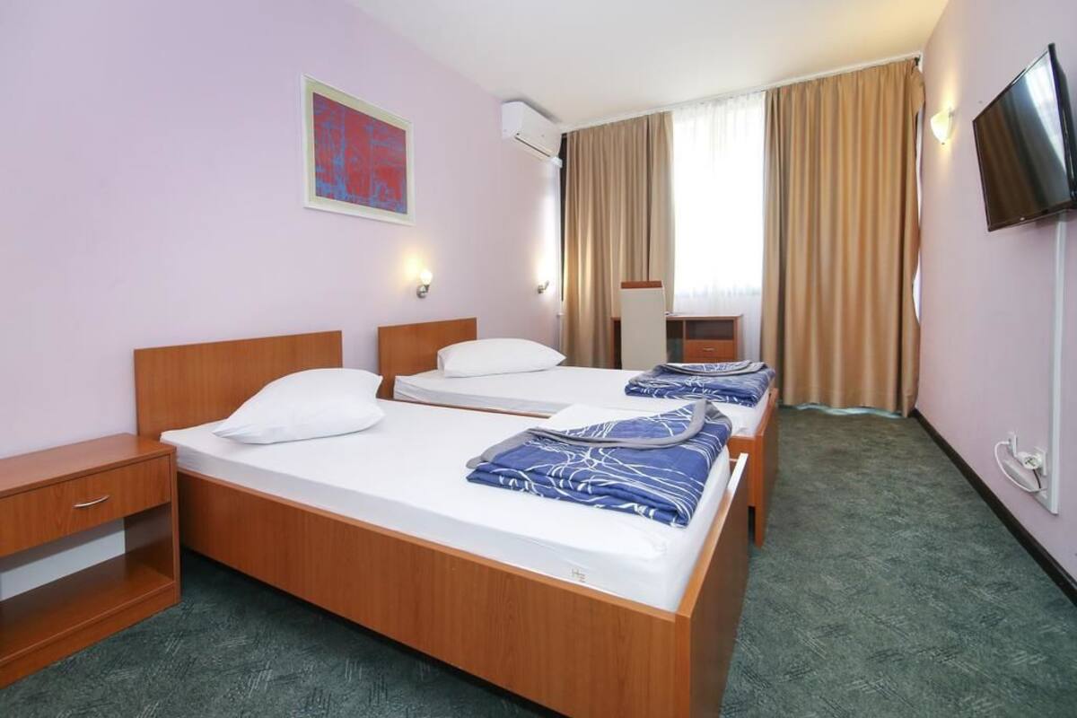 A hotel room with two single beds, a wooden nightstand, a small desk, a TV mounted on the wall, and a window with curtains.