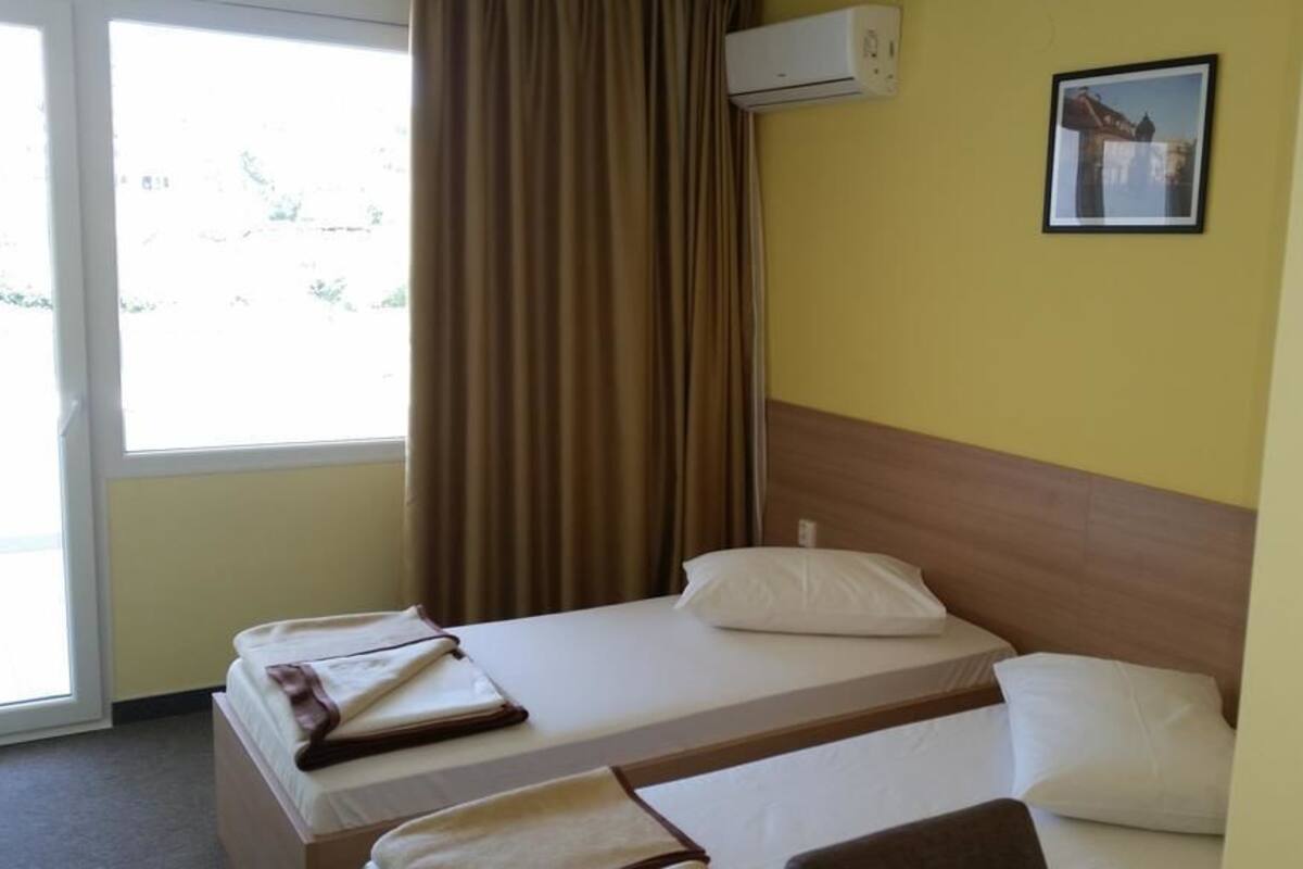 A hotel room with two beds, a window with curtains, an air conditioning unit, and a framed picture on the wall.