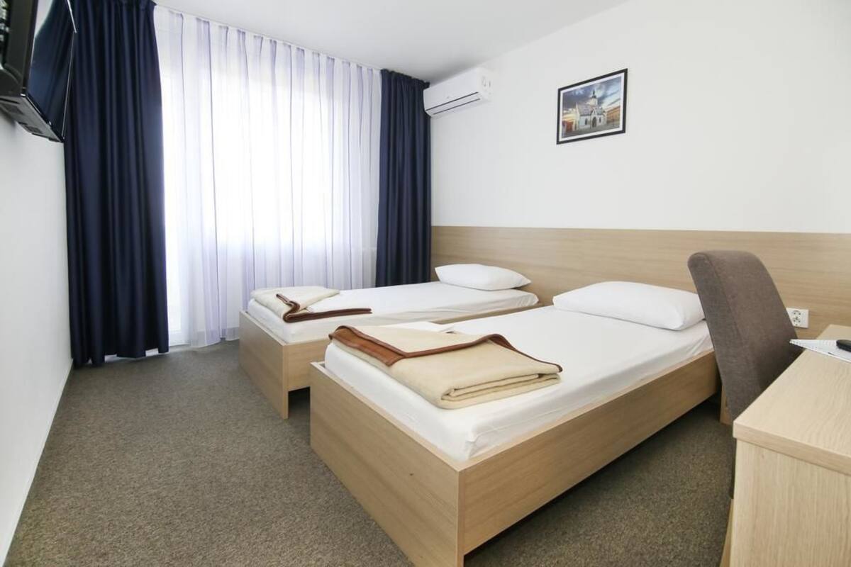 A hotel room with two single beds, a desk, a chair, and a large window with curtains.