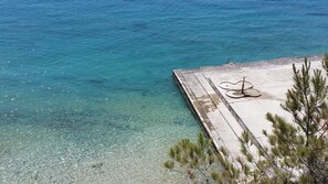 A clear view of a beach with a wooden pier extending into the turquoise sea, a bicycle resting on the pier, and sparse pine trees in the foreground. - Hotel Zagreb (Split)