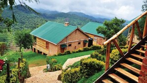 A rustic wooden house with a green roof, surrounded by lush greenery and mountains in the background. - Rihuja Safari Lodge (Bwindi)