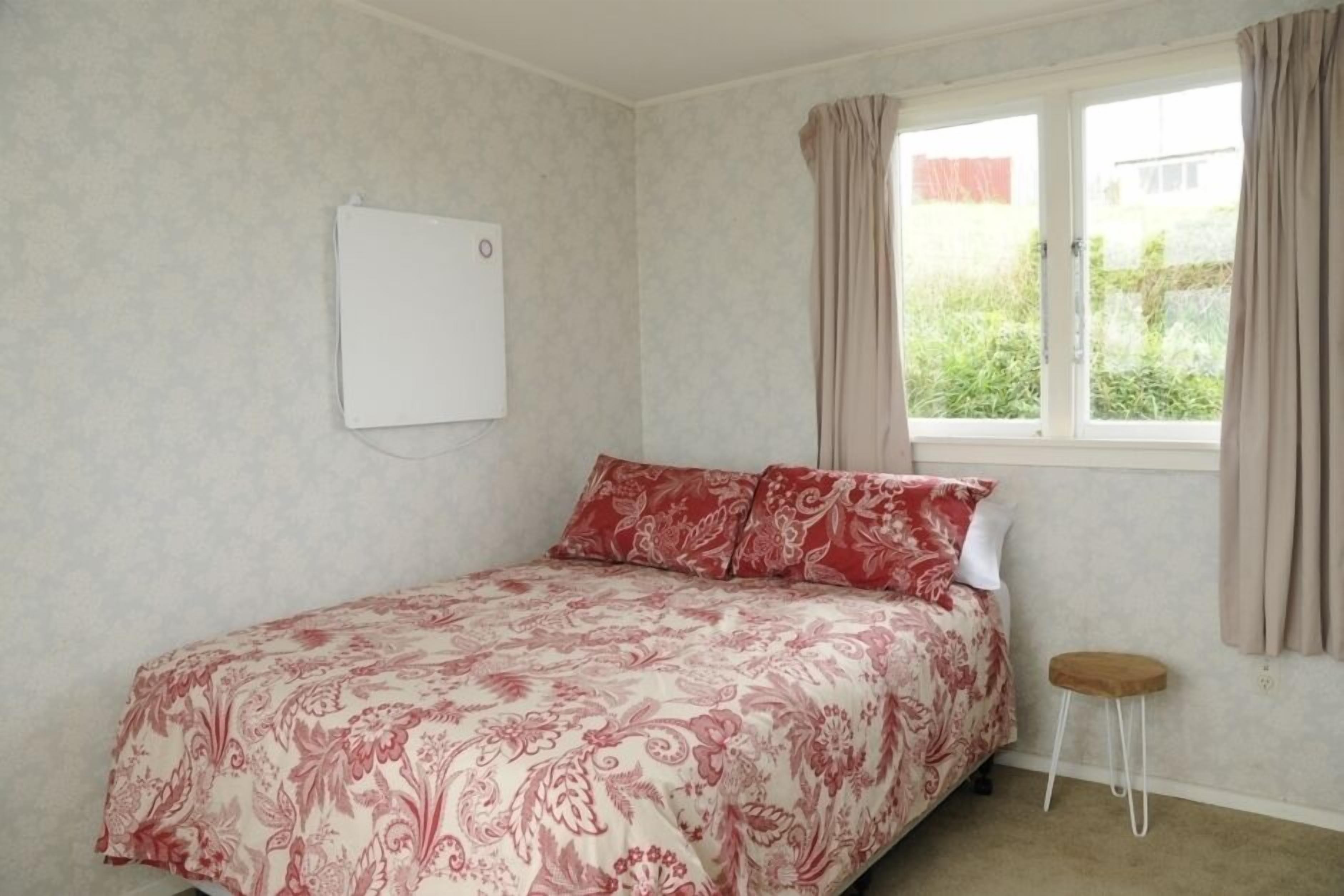 A bedroom with a bed, a small stool, a window with curtains, and a whiteboard on the wall.