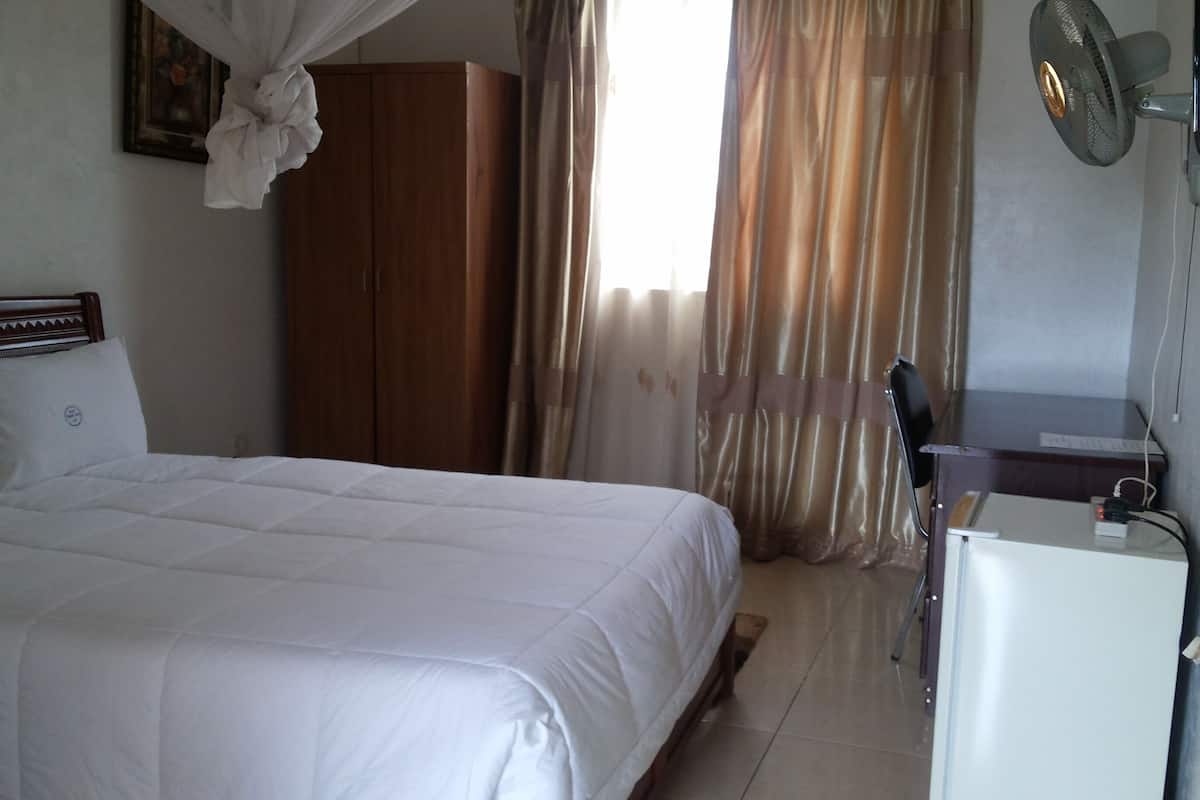Un dormitorio con cama, armario, escritorio con silla y un ventilador de pie.