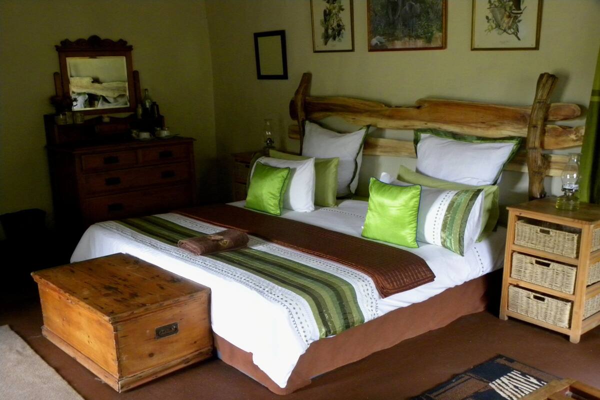 A bedroom with a wooden headboard, a wooden chest of drawers, a bed with green and white bedding, and a nightstand with a lamp.