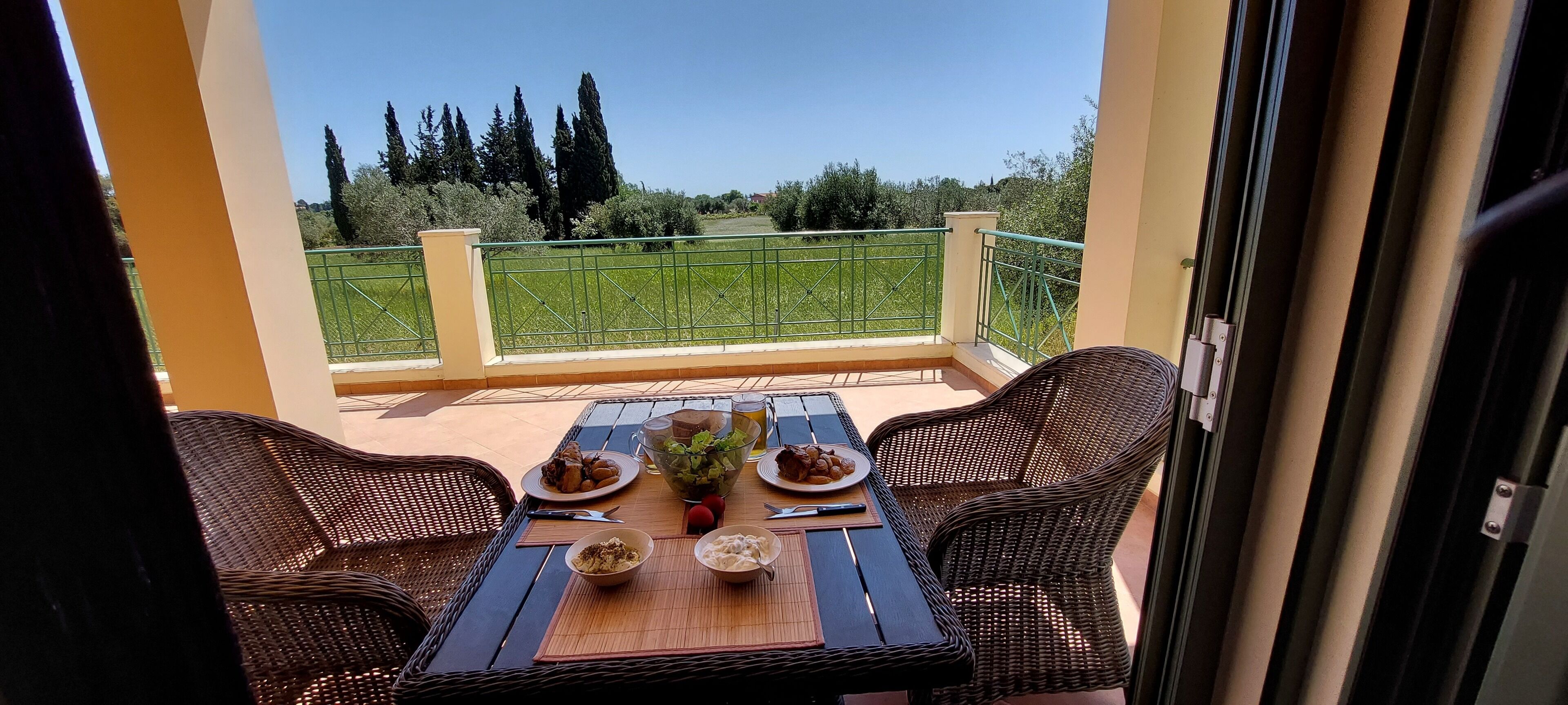 A balcony with wicker furniture, a table set with plates of food, and a view of a green field and trees.