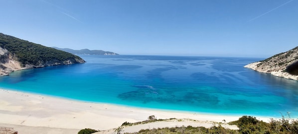 A coastal landscape with a clear blue sea, sandy beach, and green hills.