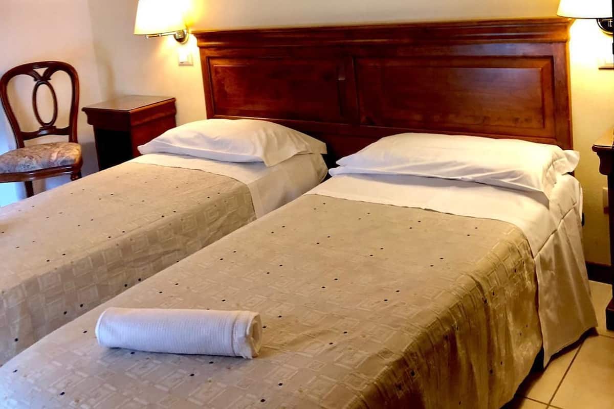 A hotel room with two single beds, a wooden headboard, a framed landscape picture on the wall, and two bedside lamps.