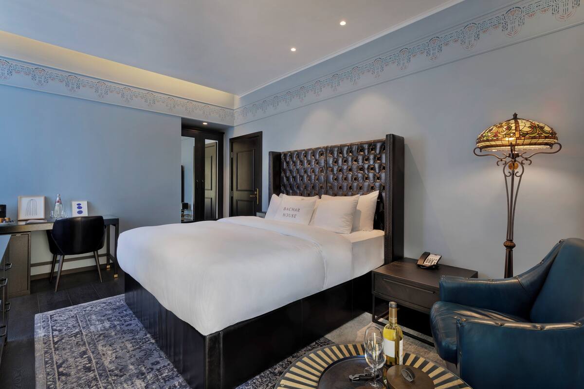 A modern hotel room with a large bed, a desk, a chair, a lamp, and a small table with a bottle and glass.
