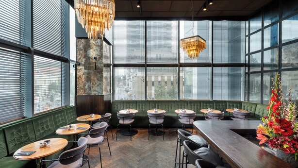 A restaurant with a high ceiling, large windows, and a mix of round and rectangular tables. - Bachar House - FATTAL COLORS (Tel Aviv)
