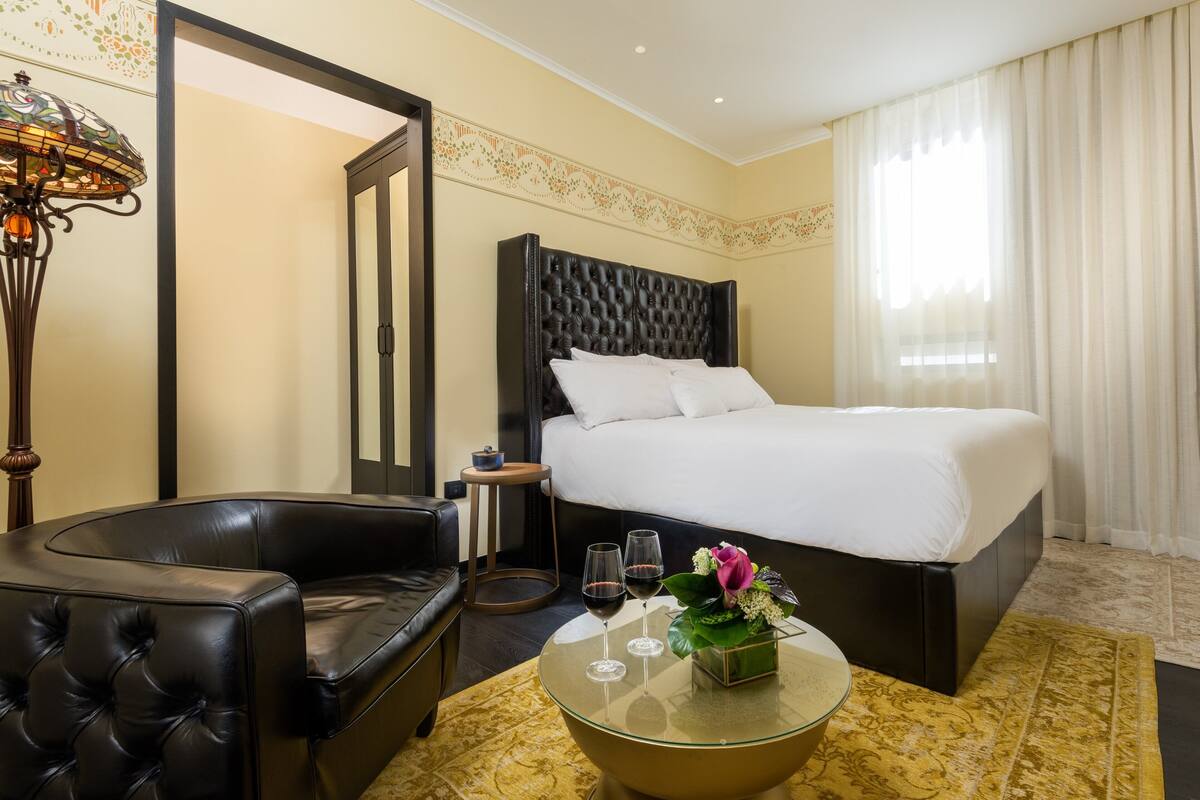 A hotel room with a large bed, a leather sofa, a glass coffee table with wine glasses and flowers, and a decorative wall panel.