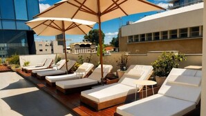 A rooftop lounge area with sunbeds and umbrellas, surrounded by buildings. - Bachar House - FATTAL COLORS (Tel Aviv)