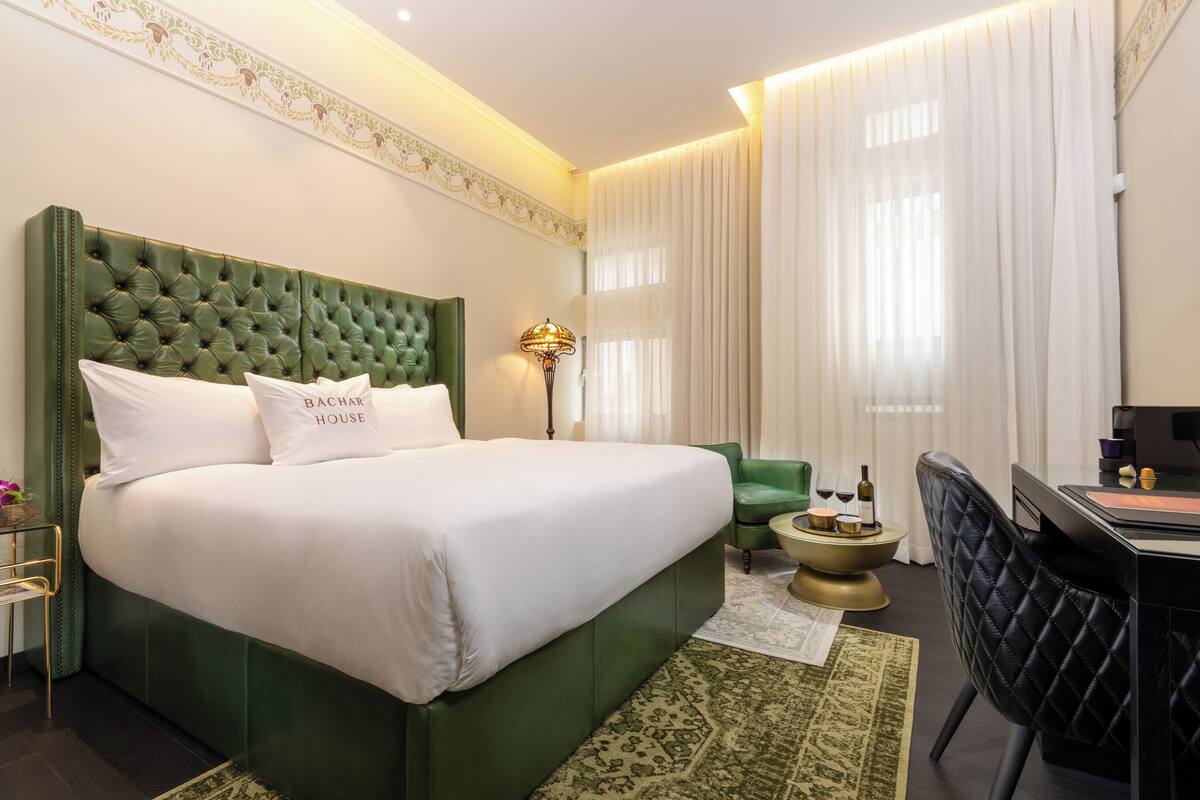 A hotel room with a green upholstered bed, a desk, a chair, and a small side table with a lamp.