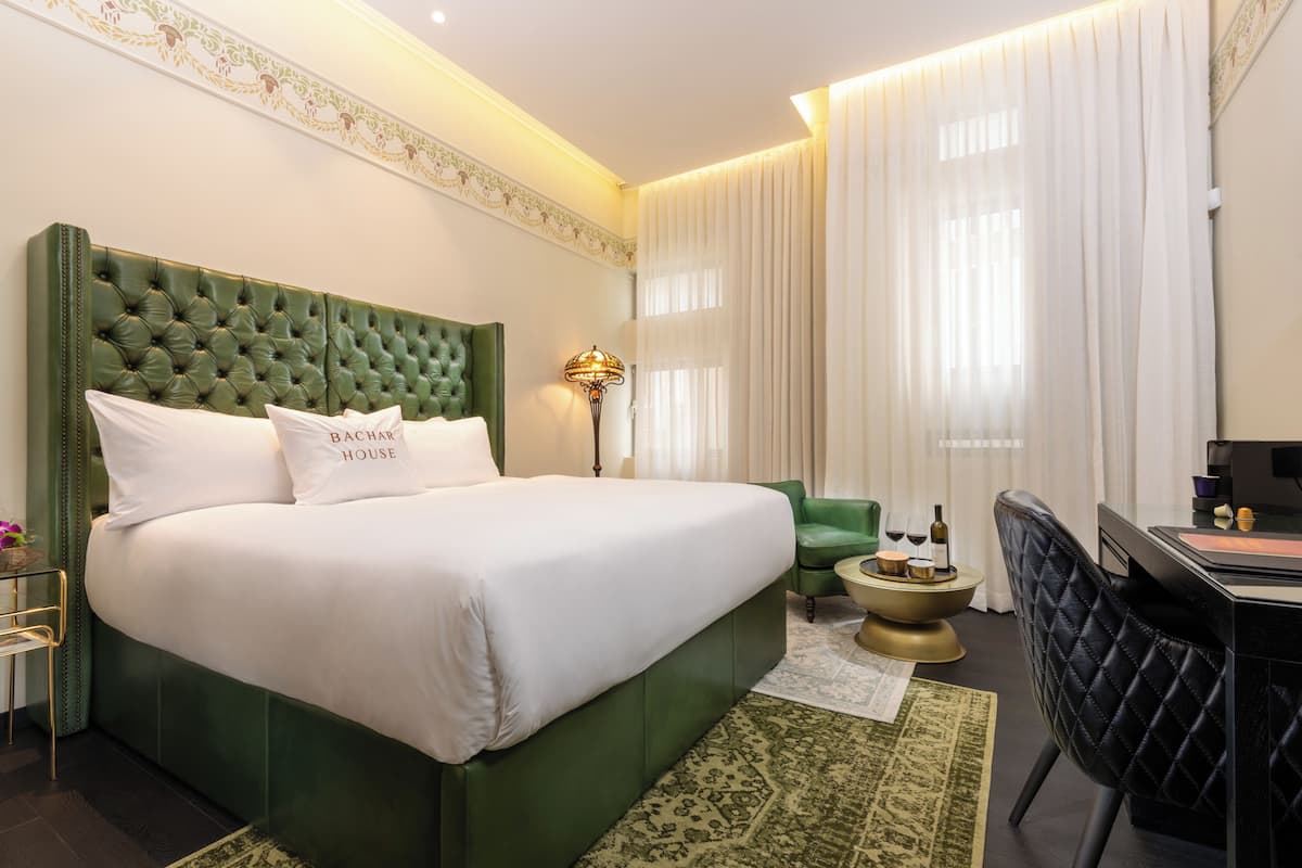 Ein Hotelzimmer mit einem grünen möblierten Bett, einem Schreibtisch, einem Stuhl und einem kleinen Beistelltisch mit einer Lampe.