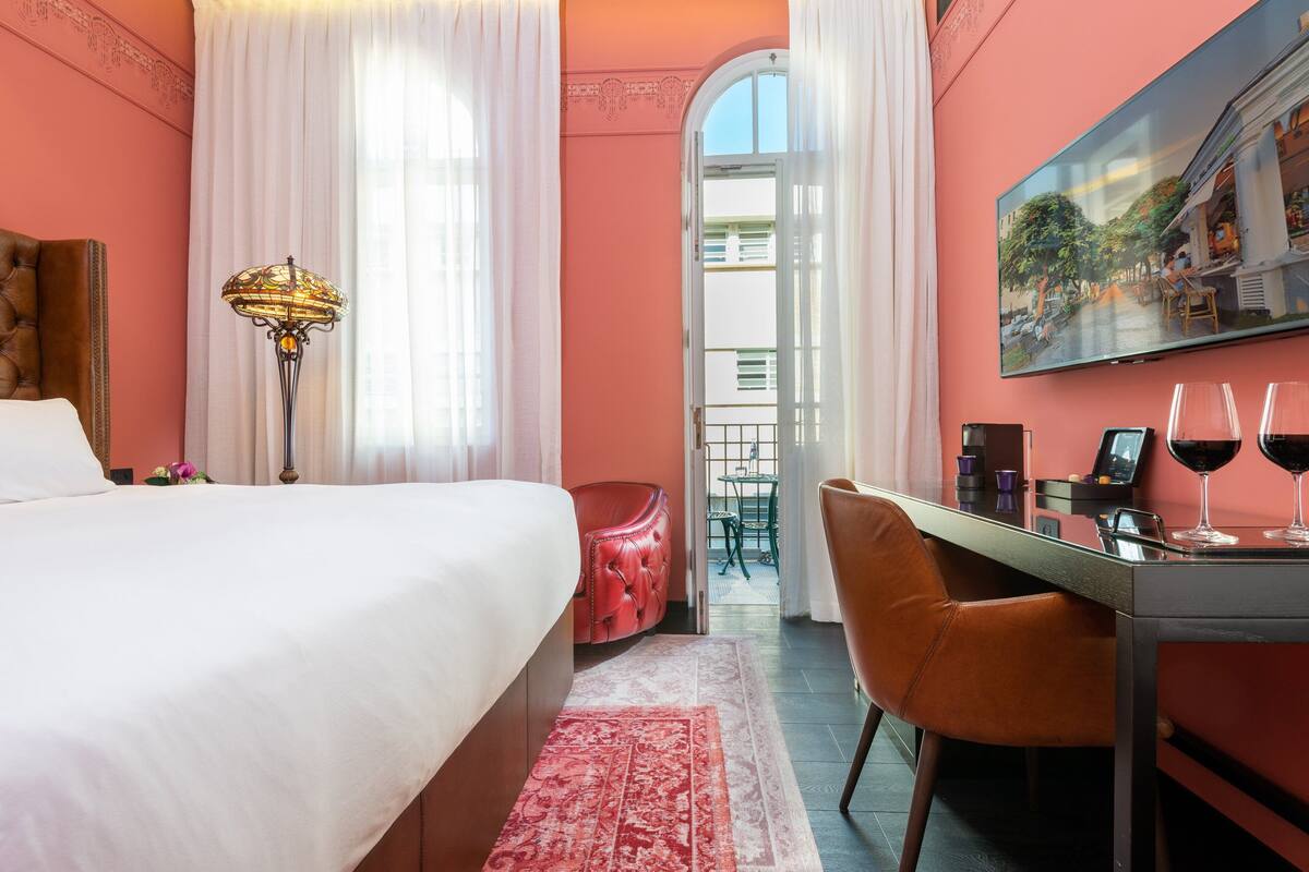 A hotel room with a bed, a desk with a computer, a chair, a lamp, and a painting on the wall.