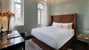 Habitación de hotel con una cama grande, un escritorio y una silla. Se ve una ventana con cortinas, una lámpara y una mesita de noche con superficie de vidrio.