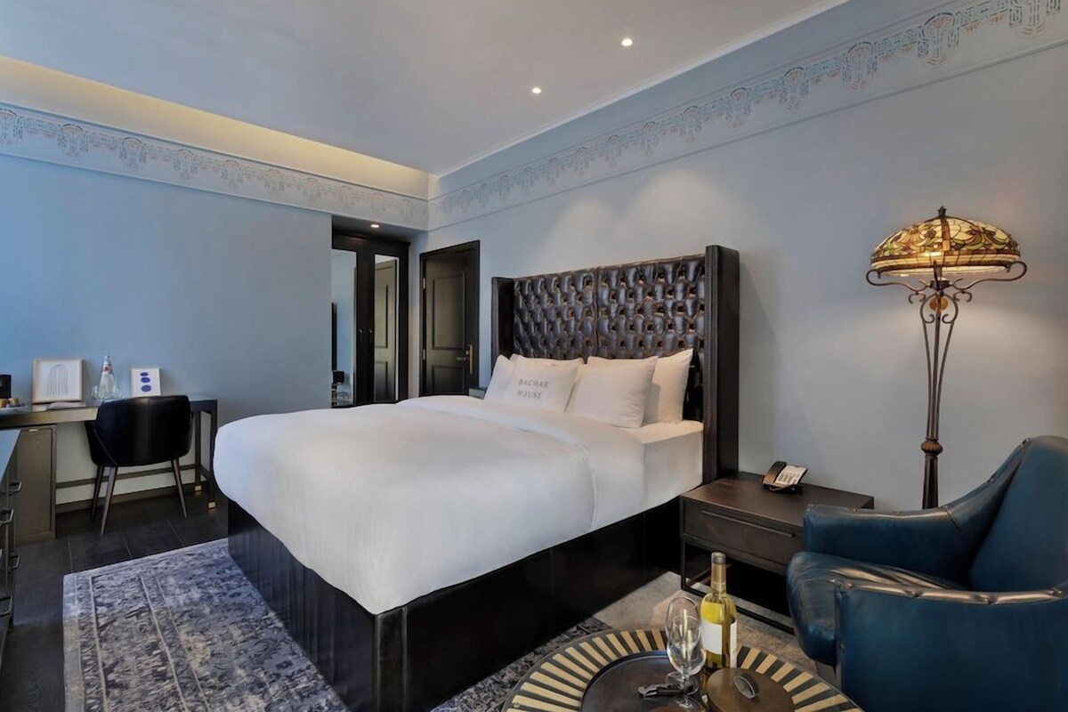 Habitación de hotel con una cama grande, un escritorio, una silla, una lámpara y una pequeña mesa con una botella y un vaso.