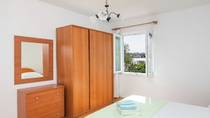 A bedroom with a wooden wardrobe, a bedside table, a bed with a green bedspread, and a window with a view of greenery and buildings.