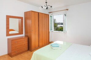 Ein Schlafzimmer mit Holzschrank, Nachttisch, einem Bett mit grüner Bettdecke und einem Fenster mit Blick auf Grünflächen und Gebäude.
