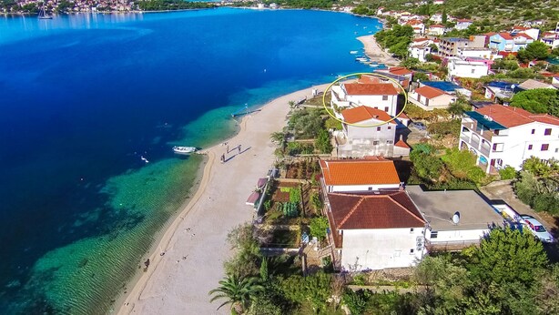 A coastal town with a sandy beach, clear blue waters, and residential buildings with red-tiled roofs.