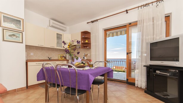 A dining area with a table set for four, a kitchen in the background, and a balcony with a TV. - Apartments Luna Gordana (Razanj)