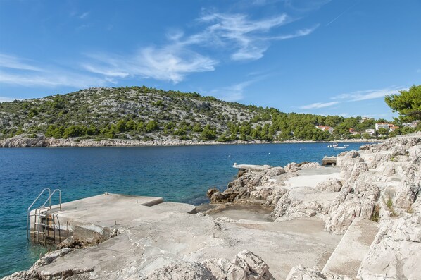 A coastal scene with a rocky pier extending into a calm sea, a hilly backdrop, and a clear sky.