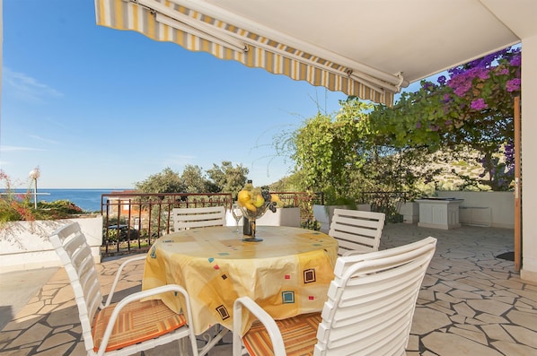 Eine Terrasse im Freien mit Tisch und Stühlen, Blick auf das Meer und einer Pergola mit Kletterpflanzen.