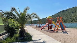 A playground with slides and swings, palm trees, and a body of water in the background.
