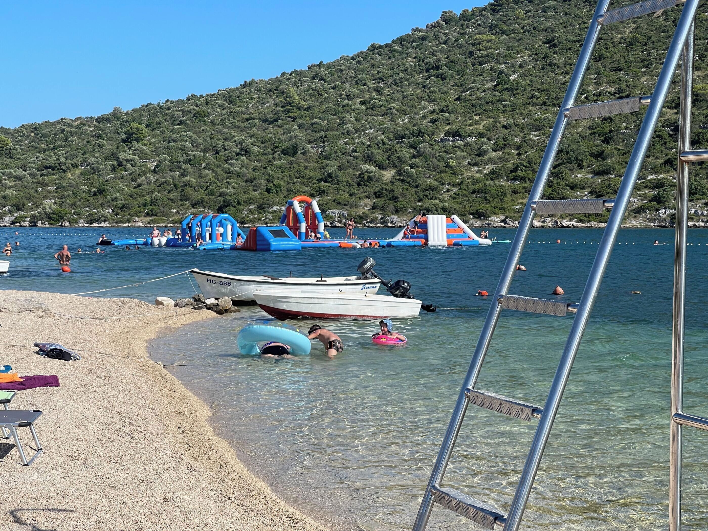 Une plage avec une eau claire et peu profonde, un bateau nommé « Resona » amarré, et des structures gonflables pour jeux aquatiques en arrière-plan.