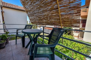 A balcony with a table and chairs, a bamboo shade, potted plants, and a view of greenery and buildings.