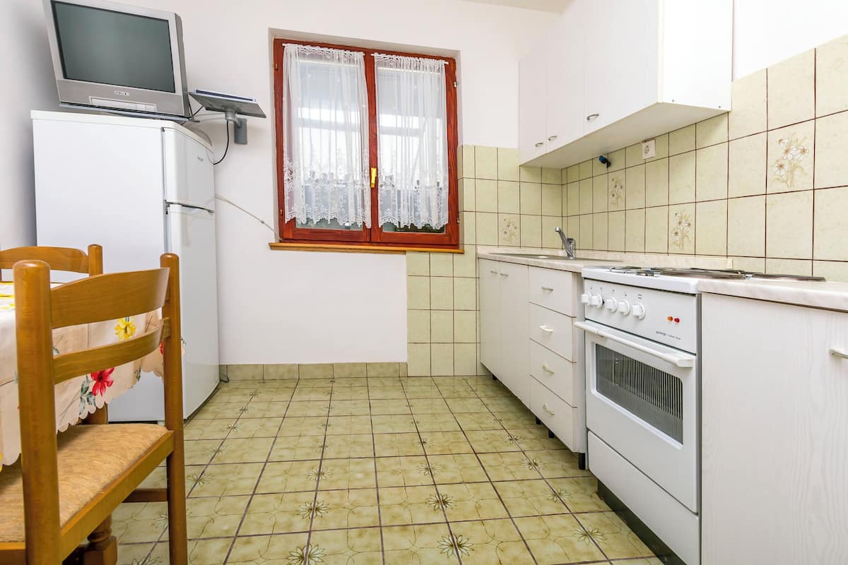 A compact kitchen with a refrigerator, oven, and a window with lace curtains.