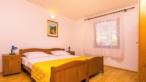 A bedroom with a wooden bed, a bedside table with a lamp, a window with sheer curtains, and a wall-mounted picture.