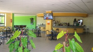 Um espaço interno amplo com várias mesas e cadeiras, um balcão com estação de bebidas e uma televisão na parede.