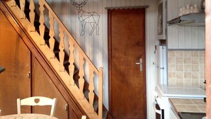 Un escalier en bois agrémenté d’un motif géométrique sur le mur, une porte en bois et un coin cuisine avec un revêtement mural en carreaux.